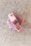 A bar of pink soap is placed on a light textured surface. The soap has small dried herbs embedded on top, giving it a natural and artisanal appearance.
