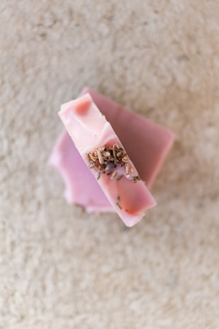 A bar of pink soap is placed on a light textured surface. The soap has small dried herbs embedded on top, giving it a natural and artisanal appearance.