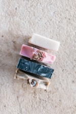 A stack of four artisanal soap bars, each with a unique color and texture, is displayed against a textured stone background. The top bar is creamy white, the second is pink with botanical elements on top, the third has a dark teal marbled pattern, and the bottom bar is a speckled tan color.