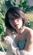 A person with short brown hair and glasses is wearing a light blue camisole and draped with a white shirt. They are standing outside with sunlight creating shadows on their face. Green foliage is in the background.