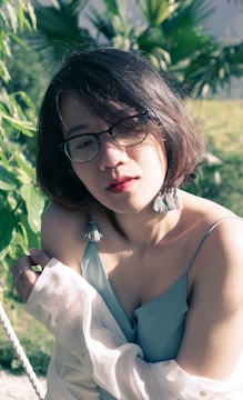 A person with short brown hair and glasses is wearing a light blue camisole and draped with a white shirt. They are standing outside with sunlight creating shadows on their face. Green foliage is in the background.