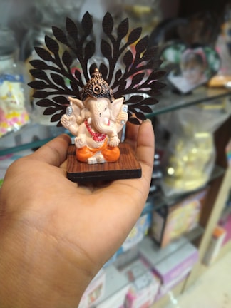 A hand holds a small statue of the Hindu deity Ganesha, which has a decorative background resembling leaves. The statue is detailed, with Ganesha seated in an ornamental headdress and wearing an orange attire. The shelf in the background holds various items, indicating that the photo was taken in a store or shop setting.