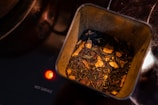 The image shows a tin container filled with loose leaf tea, which appears to be a blend containing pieces of dried fruit or spices. It is placed on a stovetop, indicated by the glowing red light and the words 'HOT SURFACE' printed nearby.