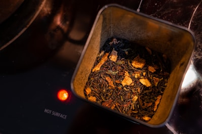 The image shows a tin container filled with loose leaf tea, which appears to be a blend containing pieces of dried fruit or spices. It is placed on a stovetop, indicated by the glowing red light and the words 'HOT SURFACE' printed nearby.