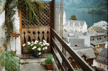 A charming balcony with a wooden lattice and lush greenery. A flowering plant in a blue pot adds color to the scene, with a background view of quaint rooftops and a serene lake surrounded by mountains.