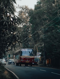A red bus with 'KSRTC' signage is driving on a tree-lined road, accompanied by a few cars in an urban setting. The background features dense greenery and overhead electric cables. Posters and signs are visible along the roadside.