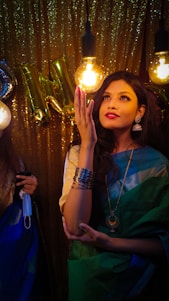 A woman in a colorful saree stands under warm lighting, admiring a bulb with an expression of wonder. She wears traditional jewelry, including earrings and bangles. The background features shiny gold streamers and balloon letters.