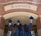 Three individuals in graduation gowns are celebrating by holding up their caps in front of a building with a brick facade. The word 'ACADEMICS' is prominently displayed above them.
