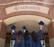 Three individuals in graduation gowns are celebrating by holding up their caps in front of a building with a brick facade. The word 'ACADEMICS' is prominently displayed above them.