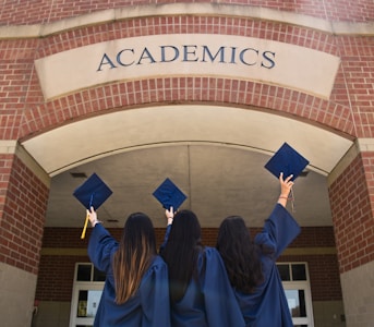 Three individuals in graduation gowns are celebrating by holding up their caps in front of a building with a brick facade. The word 'ACADEMICS' is prominently displayed above them.