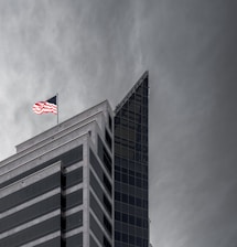 A modern building with a sharp, angular design featuring glass and concrete elements. An American flag is prominently displayed at the top, fluttering against a cloudy sky.