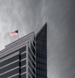 A modern building with a sharp, angular design featuring glass and concrete elements. An American flag is prominently displayed at the top, fluttering against a cloudy sky.