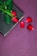 A hardcover book lies on a purple surface with a single red rose placed on top of it. The rose has vibrant green leaves, and a portion of the book’s spine is visible. Beside the book, several rose petals are scattered.