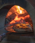 A pizza is being baked inside a wood-fired oven with visible flames and embers glowing in the background. The pizza appears to have a golden-brown crust with a slightly melted cheese and toppings.