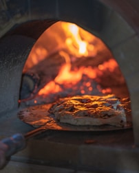 A pizza is being baked inside a wood-fired oven with visible flames and embers glowing in the background. The pizza appears to have a golden-brown crust with a slightly melted cheese and toppings.