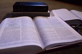 An open book with text is prominently displayed in the foreground, while another book is partially visible on the right. A closed book placed on its spine is in the background, creating a layered composition of reading materials.