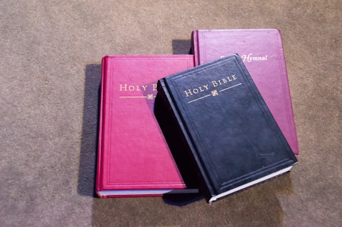 Three books are lying on a textured surface. Two of the books have red covers, one labeled 'Holy Bible' and the other 'Hymnal.' The third book, which overlaps the others, has a black cover and is also labeled 'Holy Bible.'