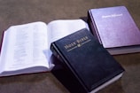 Three books are arranged on a flat surface. The first is an open book revealing pages of printed text. The second book, placed prominently in the foreground, is a closed Holy Bible with a dark cover and gold lettering. The third book is a closed Baptist Hymnal with a purple cover, displaying its title in gold lettering.