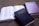 Three books are arranged on a flat surface. The first is an open book revealing pages of printed text. The second book, placed prominently in the foreground, is a closed Holy Bible with a dark cover and gold lettering. The third book is a closed Baptist Hymnal with a purple cover, displaying its title in gold lettering.