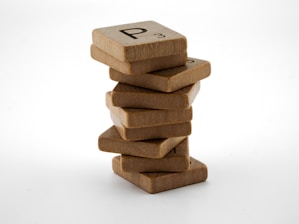 A stack of wooden game tiles, similar to Scrabble tiles, is precariously balanced against a plain white background. The tiles appear worn and some have visible letters on them.