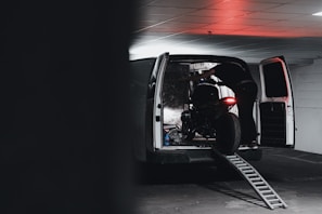 A person is loading or unloading a motorcycle into a van using a ramp in an indoor parking area. The van's rear doors are open, and there's a dim red light casting a glow on the scene.