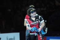 A person wearing a red and black sports shirt and a visor is standing on a dimly lit stage or arena. They are holding a blue archery bow and appear to be involved in a competitive event.