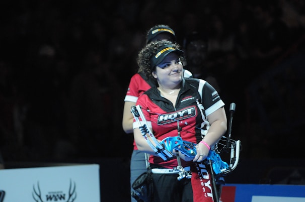 A person wearing a red and black sports shirt and a visor is standing on a dimly lit stage or arena. They are holding a blue archery bow and appear to be involved in a competitive event.