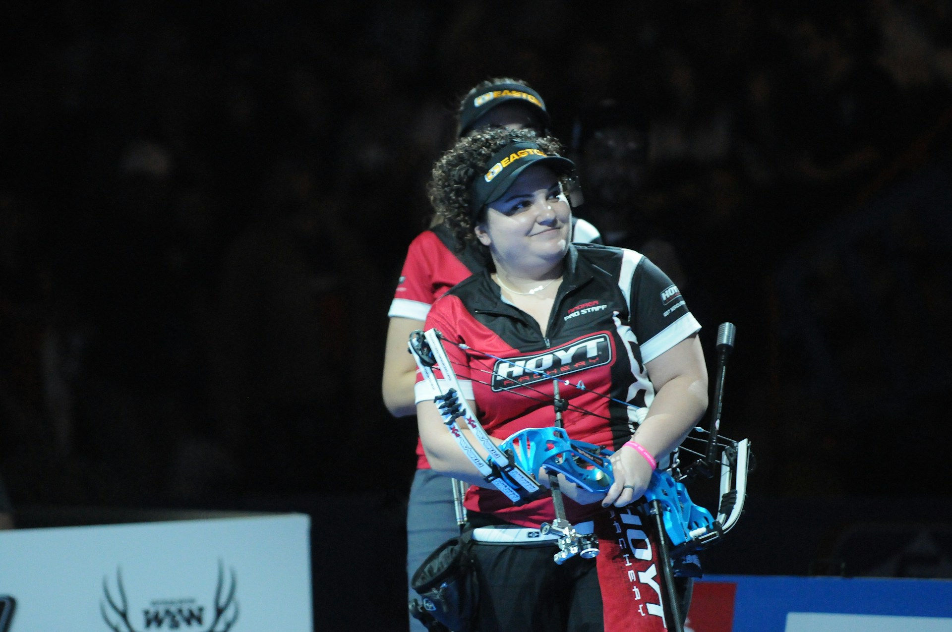 A person wearing a red and black sports shirt and a visor is standing on a dimly lit stage or arena. They are holding a blue archery bow and appear to be involved in a competitive event.