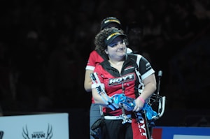 A person wearing a red and black sports shirt and a visor is standing on a dimly lit stage or arena. They are holding a blue archery bow and appear to be involved in a competitive event.