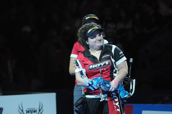 A person wearing a red and black sports shirt and a visor is standing on a dimly lit stage or arena. They are holding a blue archery bow and appear to be involved in a competitive event.