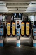A series of turnstiles at a train or metro station entrance with prominent signs reminding people to wear masks. The turnstiles feature contactless payment options and display messages about fare requirements.