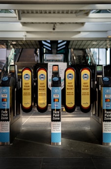 A series of turnstiles at a train or metro station entrance with prominent signs reminding people to wear masks. The turnstiles feature contactless payment options and display messages about fare requirements.