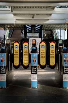 A series of turnstiles at a train or metro station entrance with prominent signs reminding people to wear masks. The turnstiles feature contactless payment options and display messages about fare requirements.