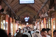 A bustling indoor market filled with people walking through a long, narrow corridor. The arched ceiling is made of brick and lined with various stalls displaying merchandise in glass cases. Overhead are electronic signs with text, and the shops have wooden elements and lighting that create a warm ambiance.