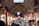 A bustling indoor market filled with people walking through a long, narrow corridor. The arched ceiling is made of brick and lined with various stalls displaying merchandise in glass cases. Overhead are electronic signs with text, and the shops have wooden elements and lighting that create a warm ambiance.