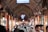 A bustling indoor market filled with people walking through a long, narrow corridor. The arched ceiling is made of brick and lined with various stalls displaying merchandise in glass cases. Overhead are electronic signs with text, and the shops have wooden elements and lighting that create a warm ambiance.