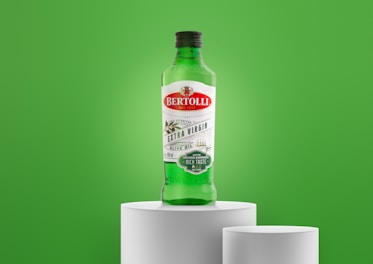 A green bottle of Bertolli Extra Virgin Olive Oil is placed on a white pedestal against a vibrant green background. The label features red and green colors, indicating it is cold extracted and suitable for dressings and marinades.