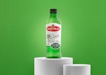 A green bottle of Bertolli Extra Virgin Olive Oil is placed on a white pedestal against a vibrant green background. The label features red and green colors, indicating it is cold extracted and suitable for dressings and marinades.