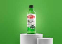 A green bottle of Bertolli Extra Virgin Olive Oil is placed on a white pedestal against a vibrant green background. The label features red and green colors, indicating it is cold extracted and suitable for dressings and marinades.