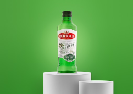 A green bottle of Bertolli Extra Virgin Olive Oil is placed on a white pedestal against a vibrant green background. The label features red and green colors, indicating it is cold extracted and suitable for dressings and marinades.