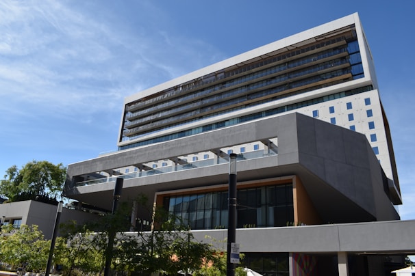 A modern, multi-story building with a sleek architectural design featuring geometric shapes and clean lines. The facade includes large glass windows and balconies, and it is surrounded by trees and clear blue sky.