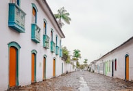 A picturesque cobblestone street lined with historic white colonial-style buildings adorned with vibrant doors and windows. The architecture features turquoise accents and wooden shutters. Tall palm trees punctuate the scene, adding a tropical touch to the serene environment.
