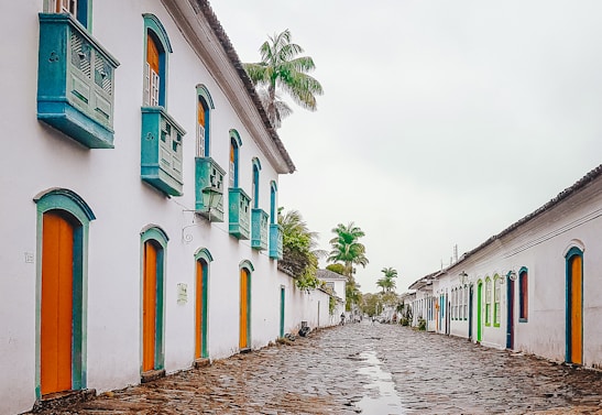 A picturesque cobblestone street lined with historic white colonial-style buildings adorned with vibrant doors and windows. The architecture features turquoise accents and wooden shutters. Tall palm trees punctuate the scene, adding a tropical touch to the serene environment.