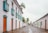 A picturesque cobblestone street lined with historic white colonial-style buildings adorned with vibrant doors and windows. The architecture features turquoise accents and wooden shutters. Tall palm trees punctuate the scene, adding a tropical touch to the serene environment.