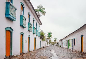 A picturesque cobblestone street lined with historic white colonial-style buildings adorned with vibrant doors and windows. The architecture features turquoise accents and wooden shutters. Tall palm trees punctuate the scene, adding a tropical touch to the serene environment.