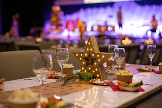A festive dining setup features a large, wooden star centerpiece accented with small lights, surrounded by glasses, dishes, and decorative greenery on a table. In the background, colorful, blurred lights create a vibrant ambiance.
