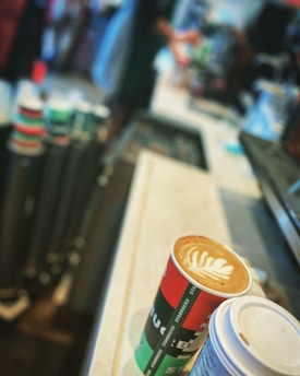 Two cups of coffee are placed on a countertop, with the focus on a latte decorated with latte art. In the background, there are blurred images of people and a stack of disposable coffee cups.