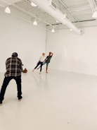In a spacious, light-filled studio with white walls and exposed beams, two people are posing playfully while a photographer captures their moment. The setting is minimalistic, emphasizing the interaction between the photographer and the subjects.