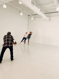 In a spacious, light-filled studio with white walls and exposed beams, two people are posing playfully while a photographer captures their moment. The setting is minimalistic, emphasizing the interaction between the photographer and the subjects.