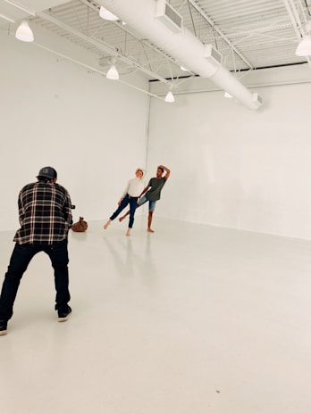 In a spacious, light-filled studio with white walls and exposed beams, two people are posing playfully while a photographer captures their moment. The setting is minimalistic, emphasizing the interaction between the photographer and the subjects.